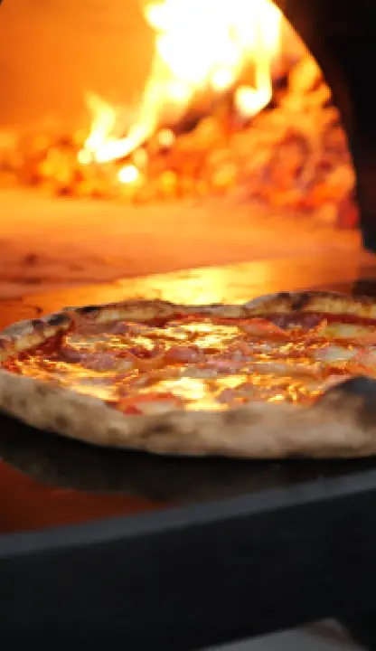 Three uncooked pizzas, topped with various ingredients including cheese and sauce, are placed inside a glowing wood-fired oven. Flames are visible in the back and on the right side of the oven.