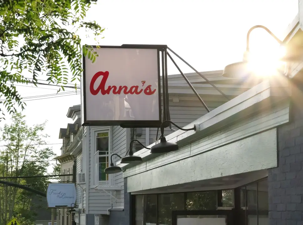 A square white sign with the name "Anna's" written in red cursive font is mounted on a building. The sun shines brightly from behind the sign, creating a lens flare and sun rays. A black industrial-style light fixture hangs to the right of the sign, and power lines are visible on the left. The sky behind is blue with scattered white clouds.