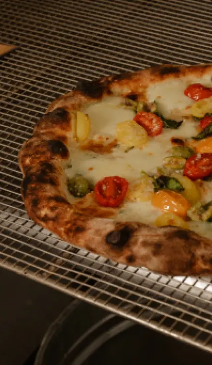 A close-up, angled shot of a Neapolitan-style pizza with a blistered, slightly charred crust, resting on a metal cooling rack. The pizza is topped with melted white cheese, sliced red and yellow cherry tomatoes, and possibly some green herbs.