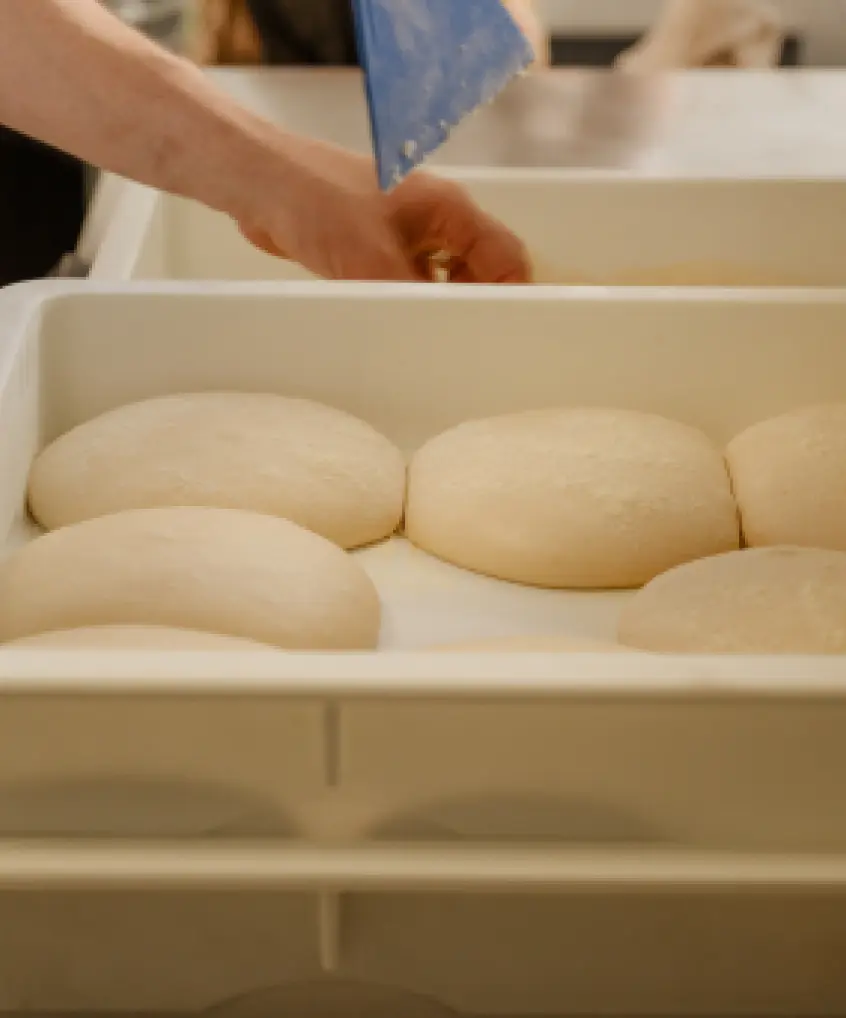 A shallow depth of field shot shows several mounds of raw pizza dough resting in white plastic proofing bins. In the blurred background, a person's hands are visible, along with a blue dough scraper, suggesting ongoing dough preparation.