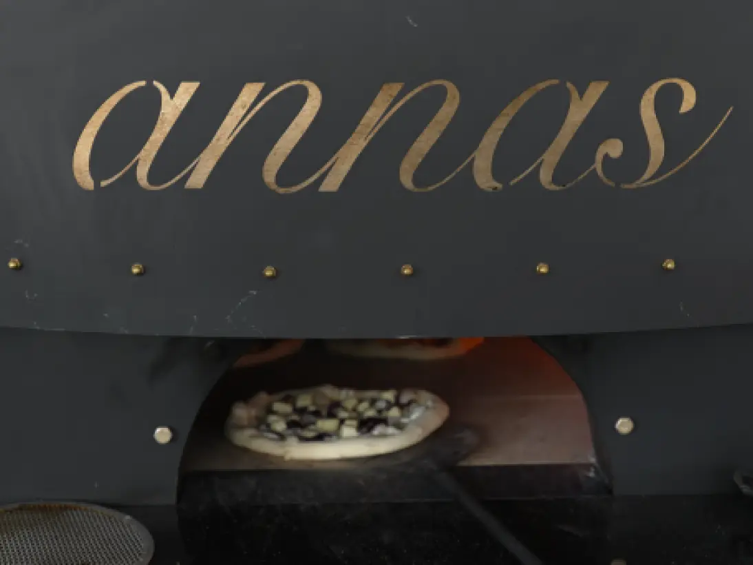 A pizza oven with "annas" written in a gold, cursive font on its dark exterior. Below the lettering, a pizza topped with what appears to be dark and light colored ingredients is being slid into the glowing interior of the oven with a pizza peel.