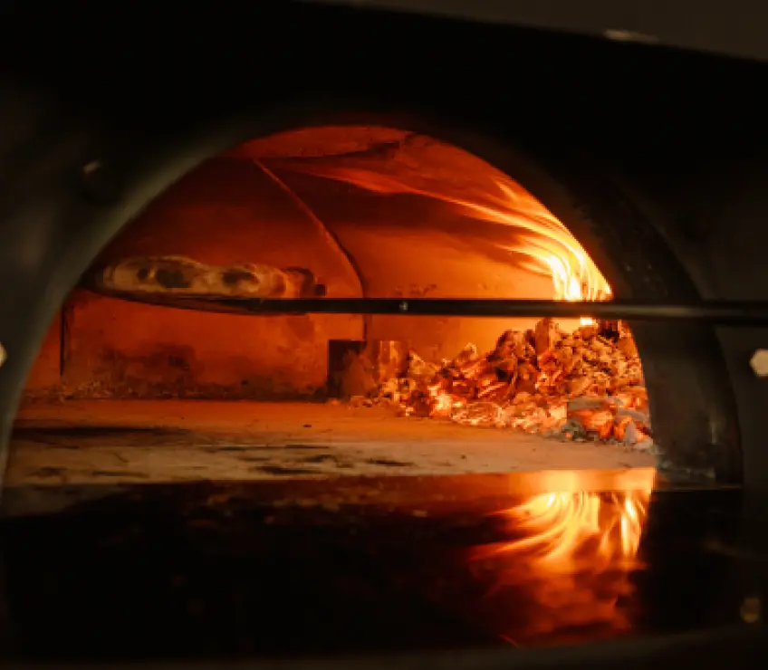 A perfectly cooked, bubbling pizza with golden-brown crust and melted cheese sits inside a wood-fired oven. Intense flames are visible in the background, illuminating the pizza