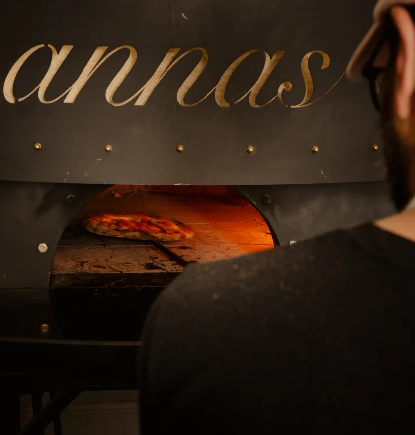 The image shows a person from behind, looking into a pizza oven. The oven is built into a dark surface with the name "annas" written in a stylized, light-colored font above the opening. Inside the oven, a pizza is being cooked, visible on a peel (a long-handled tool used to slide pizzas in and out of the oven) and glowing from the heat of the fire within. The scene suggests a cozy, possibly artisanal, pizza-making environment.