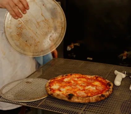 A freshly baked pizza with melted cheese and red sauce, featuring a slightly charred crust, rests on a metal cooling rack. To the left of the pizza, a round metal screen for cooling or holding pizza is visible. Above the screen, a person's hand holds a dirty, round metal pizza pan or lid. In the background, a dark surface is visible, possibly part of a kitchen area.