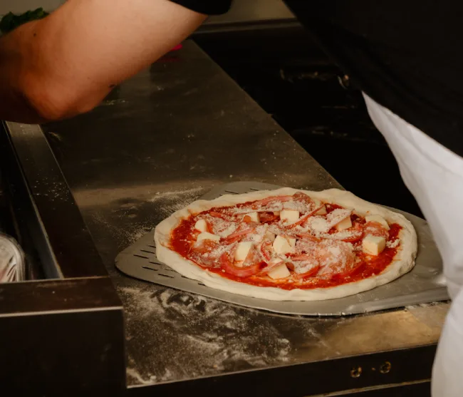 A pizza, topped with red sauce, white cheese cubes, and what appears to be sliced meat, rests on a metal pizza peel. The peel is positioned on a stainless steel counter dusted with flour. To the right, part of a person wearing a white apron is visible, and in the background, another person's arm and a dark surface can be seen.