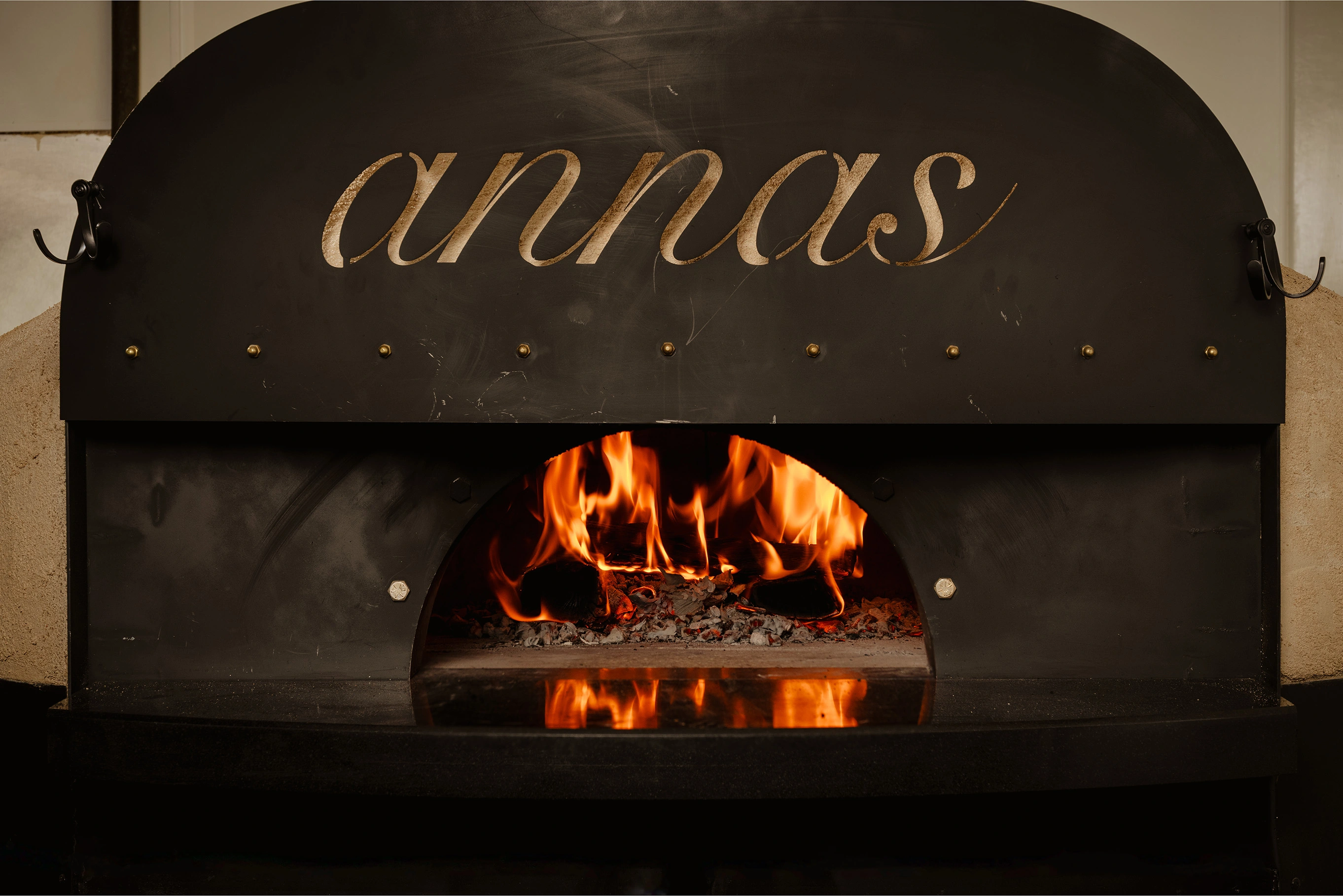 A close-up shot of a black, brick pizza oven with a blazing fire inside. The word "annas" is written in a graceful, handwritten font across the top of the oven.