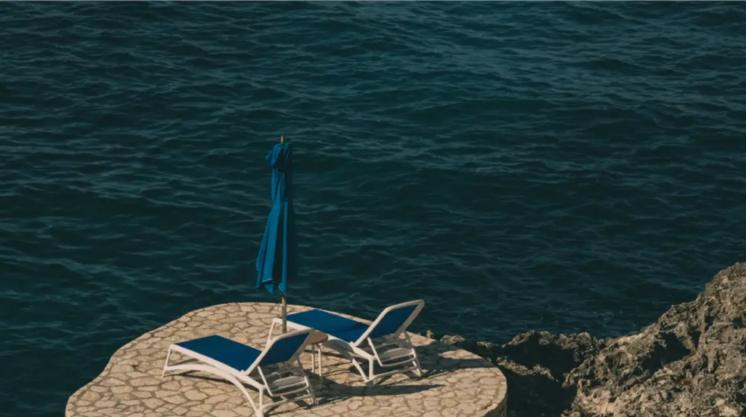 A secluded stone sun deck at Rockhouse Hotel in Negril, Jamaica, with two blue lounge chairs and a matching umbrella perched above the deep blue Caribbean Sea.